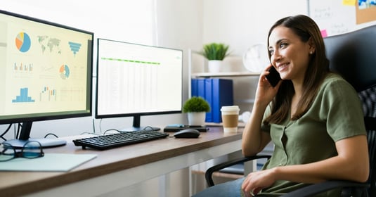 A professional woman looks at her dual monitor set-up while talking on her cell phone 
