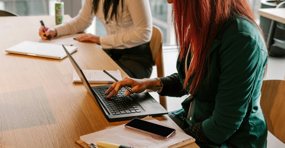 Two professional women work from laptops at a large conference table